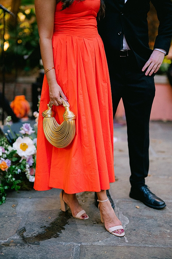 Wedding guest outfit featuring a coral wedding guest dress with gold handbag and nude strappy heels, paired with a black suit on a stone walkway with floral greenery