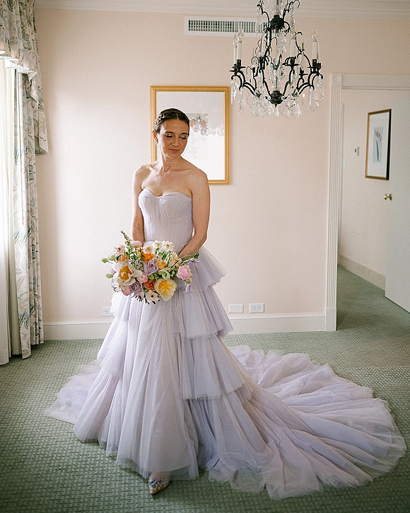 Bridal portrait in a lavender wedding dress, holding a pastel bouquet of roses and ranunculus in a window-lit room with chandelier