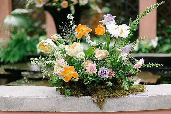 Wedding floral centerpiece with garden roses, ranunculus, poppies and peonies, set on moss atop a stone ledge by a courtyard fountain