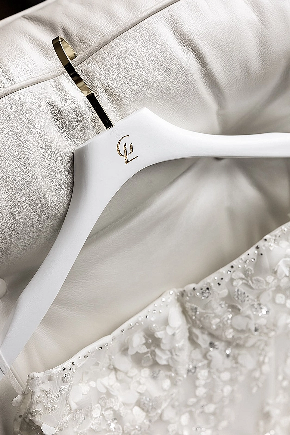 Wedding dress close-up showing lace wedding dress detail with sequins and beadwork on a monogrammed hanger, draped over a white leather chair