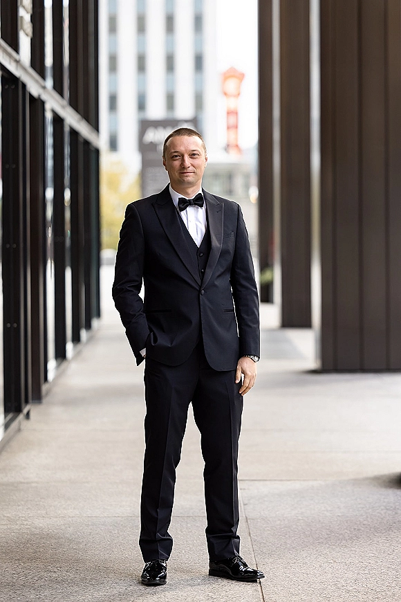 Groom portrait in a black tuxedo groom look, standing full length with hands in pockets on a city sidewalk by modern columns
