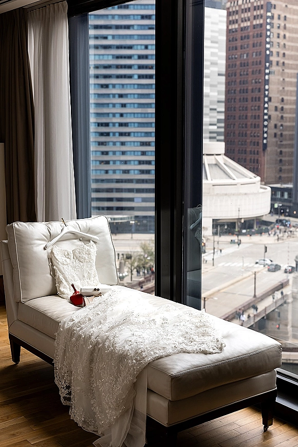 Wedding dress, lace wedding dress hanging by a floor-to-ceiling window with heels and jewelry, city skyline visible beyond hotel curtains