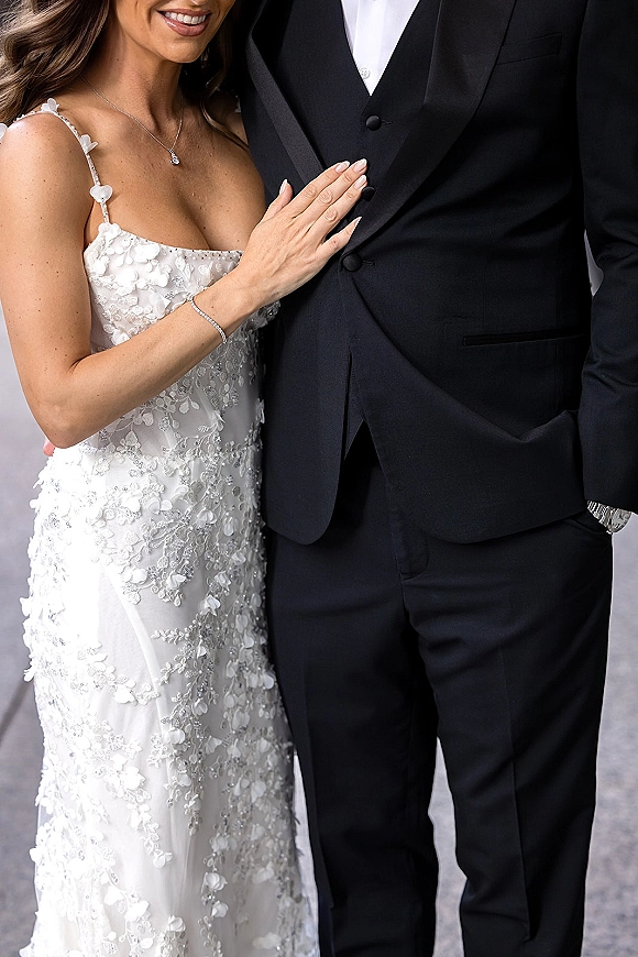 Couple portrait of bride with hand on groom’s chest, showing lace floral appliqué dress, diamond necklace, and black tuxedo on neutral backdrop