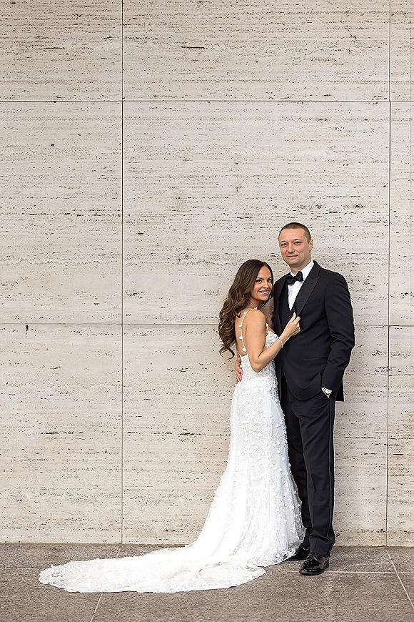 Couple portrait of bride and groom hugging, her lace gown with train beside his tuxedo and bow tie against a stone wall backdrop