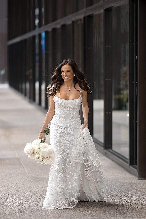 Bridal portrait of a bride holding bouquet of white flowers in a lace wedding dress, walking by a glass building on a city sidewalk