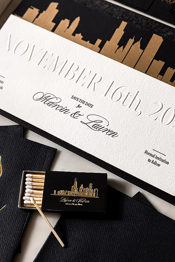Wedding invitations and save the date card in a black and white flat lay with embossed calligraphy, black liners, and gold foil skyline