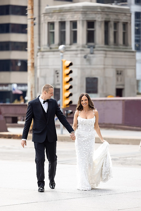 Couple portrait of bride and groom walking hand in hand on a city street, her lace wedding dress and train flowing beside his black tuxedo