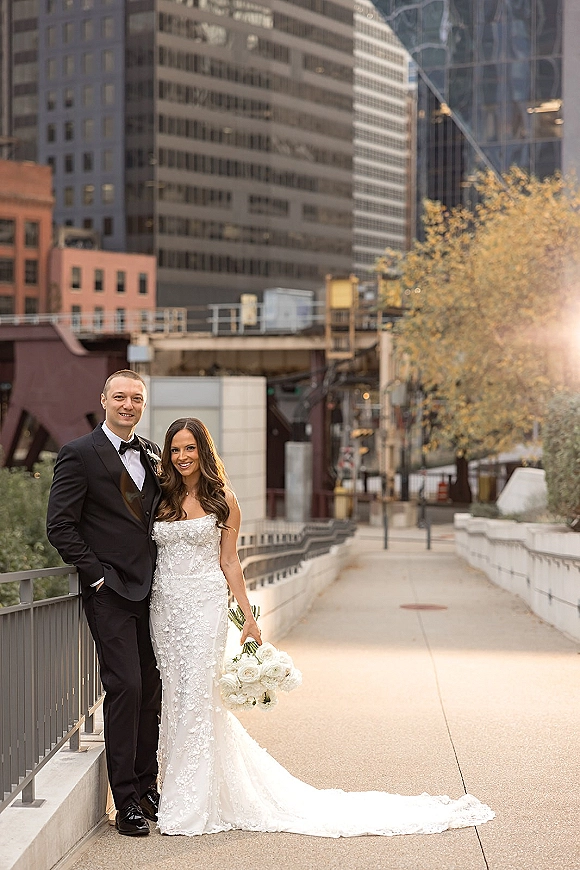 Couple portrait of bride in strapless lace gown holding white rose bouquet beside groom in tuxedo on a sunlit city bridge with flare