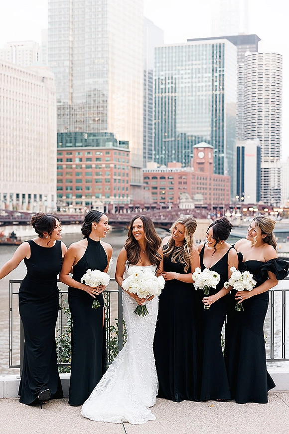 Bride with bridesmaids in black bridesmaid dresses holding white bouquets, posing by a waterfront railing with city skyline and bridge behind