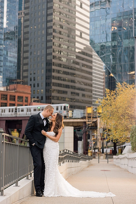 Couple portrait urban wedding portrait of bride in strapless gown with long train and groom in tuxedo leaning by railing, skyline behind
