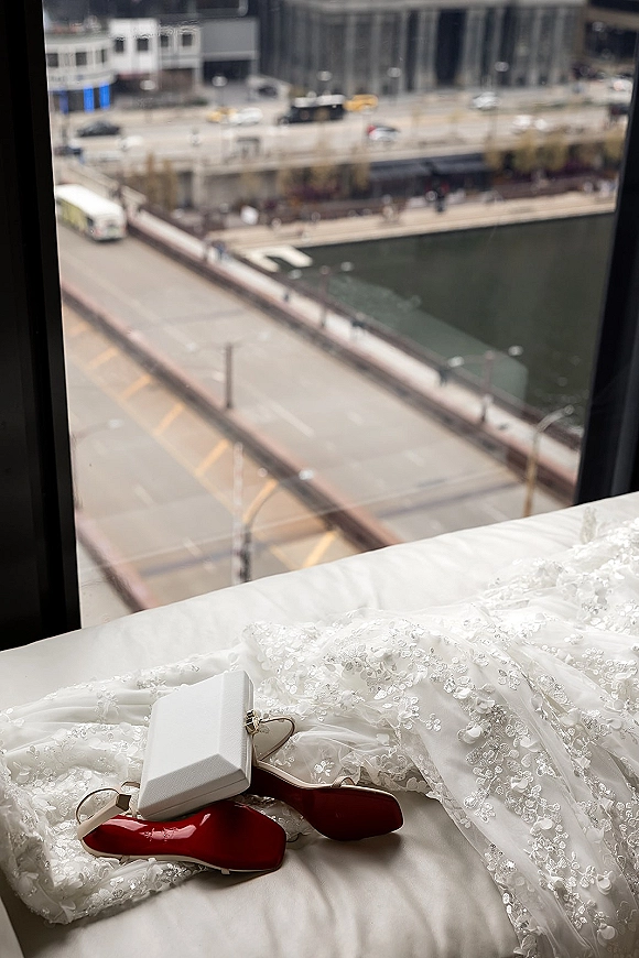 Wedding dress detail with lace wedding dress appliqué and beaded tulle, bridal heels with red soles and ring box by a window overlooking the waterfront city