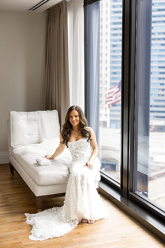 Bridal portrait of bride sitting on chaise in a strapless lace wedding dress by floor-to-ceiling hotel windows with city view
