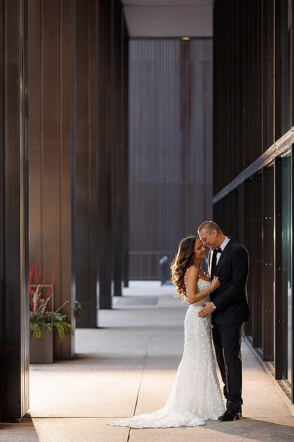 Couple portrait of bride and groom embrace, forehead touching under a modern covered walkway with columns, her lace train flowing