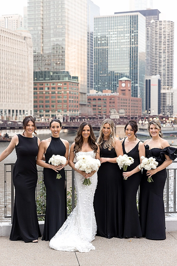 Bride with bridesmaids group photo on a waterfront railing, black dresses and white bouquets, with city skyline, bridge, and river behind