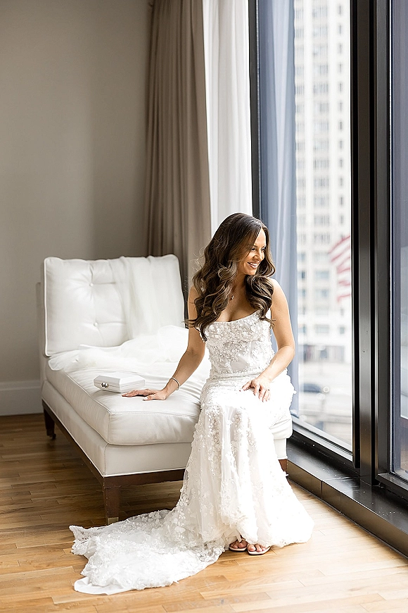 Bridal portrait of a bride sitting by window in a strapless lace gown with long train on a chaise lounge, city buildings beyond curtains