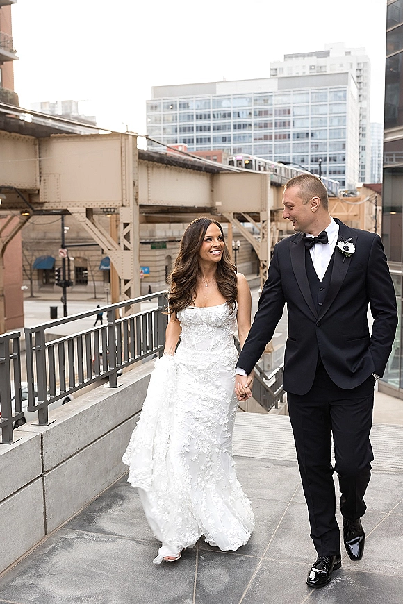 Couple portrait of bride and groom walking holding hands on a rooftop walkway with city buildings and elevated train tracks behind