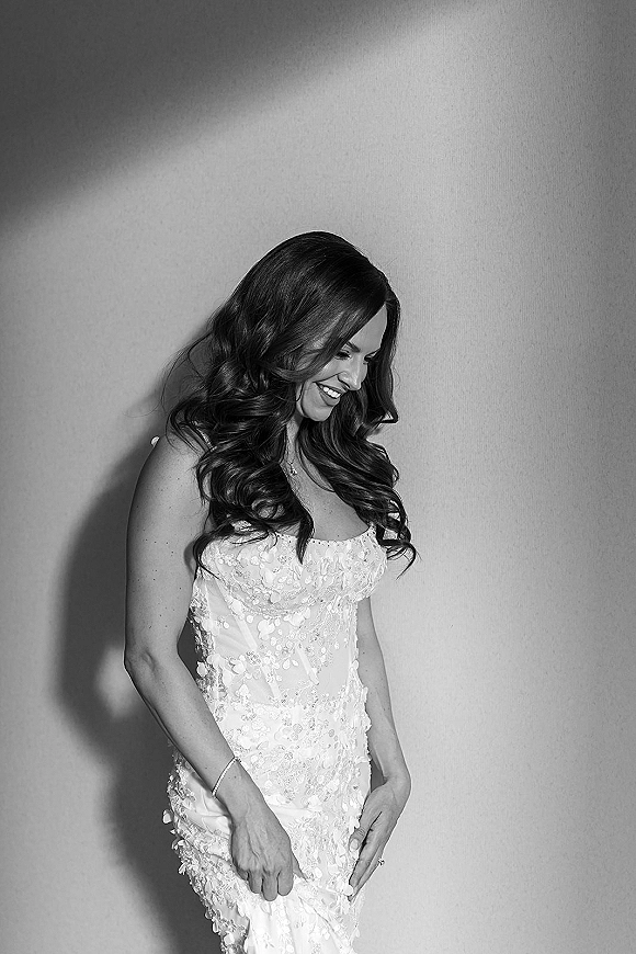 Bridal portrait in black and white of a bride looking down in a strapless lace wedding dress with bracelet, ring, and necklace by a plain wall