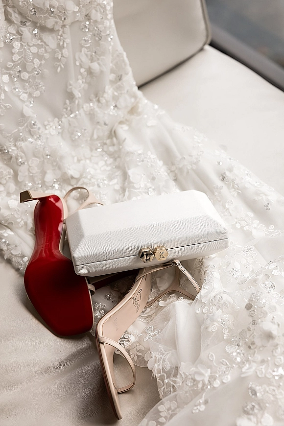 Bridal accessories flatlay with wedding dress detail, beaded lace gown and tulle, nude strappy red-sole heels, and monogram white clutch on a chair