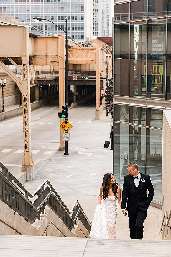 Couple portrait of bride and groom walking hand in hand down city stairs beneath elevated train tracks, bride in lace gown and groom in tuxedo