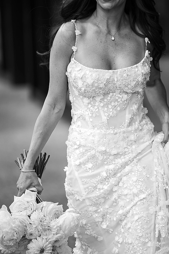Wedding dress with 3d floral appliqué and spaghetti straps, bride holding white rose bouquet and wearing pendant necklace on outdoor walkway
