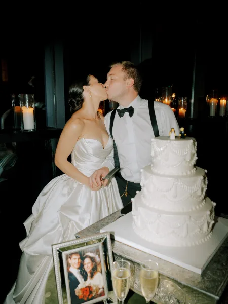Cake cutting moment as bride and groom kiss beside a three-tier wedding cake, candlelit table with champagne flutes in a dark interior