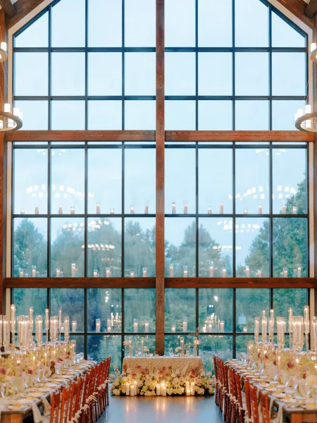 Reception tablescape with candlelit wedding reception details, floral centerpieces and taper candles on a long table by window walls at dusk