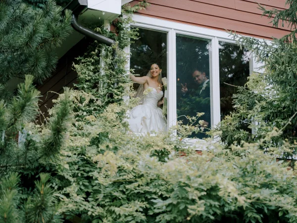 Wedding couple portrait of bride at window in a strapless satin gown and veil, groom behind glass in black suit with boutonniere at ivy-covered house