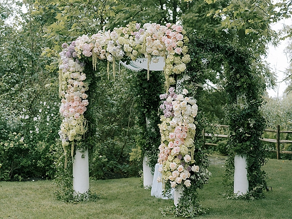 Wedding ceremony arch with roses and hydrangeas on white pillars, draped fabric and greenery garland, set on a garden lawn with trees