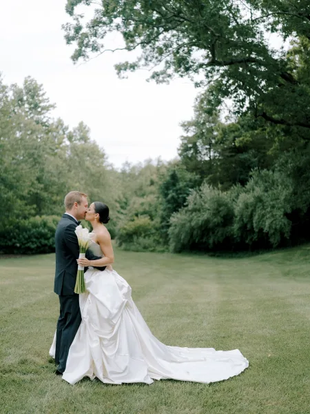 Wedding kiss portrait of bride and groom kissing outdoors, bride holding white calla lilies, long dress train on grass lawn with trees behind