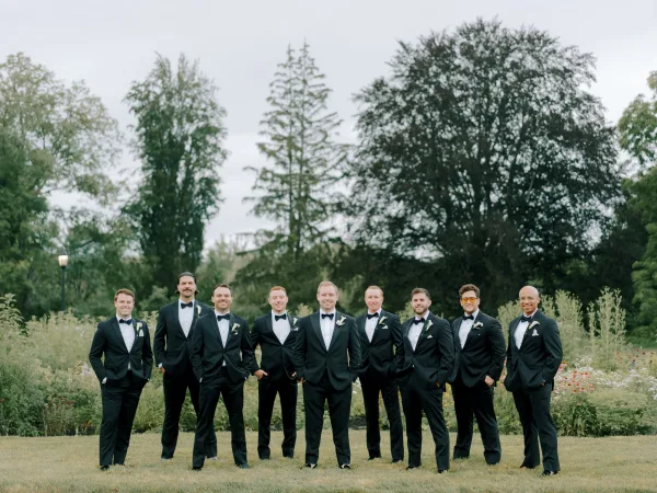 Groomsmen portrait of a lineup in black tuxedos with bow ties and sunglasses, standing on a garden lawn with wildflowers under an overcast sky