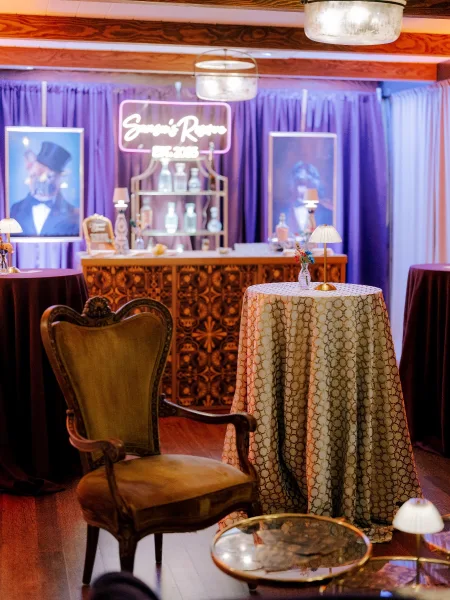 Wedding lounge decor with velvet chair, neon sign, and patterned cocktail tables beside a carved wood bar under purple uplighting
