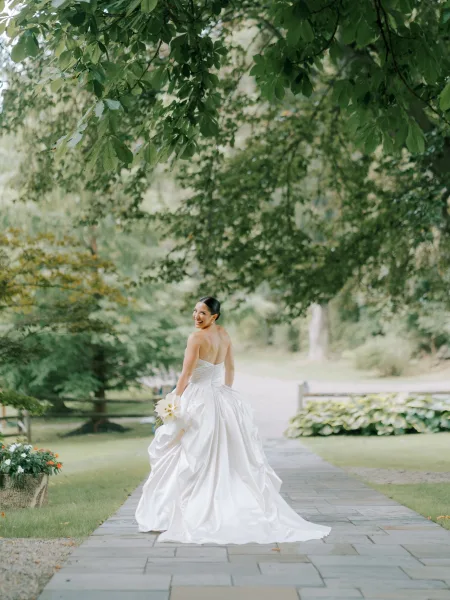 Bridal portrait of a bride in a strapless satin wedding dress looking back, holding a calla lily bouquet on a garden stone walkway