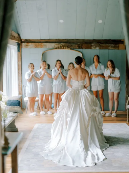 Wedding dress reveal as bride in a strapless gown with long train faces bridesmaids in pajamas in a rustic, window-lit bridal suite