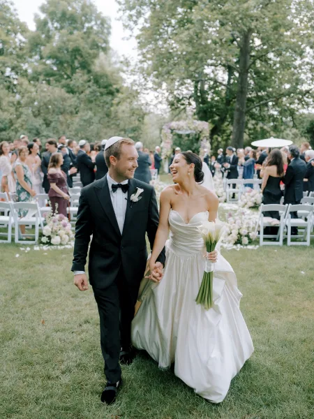 Wedding recessional as bride and groom walk hand in hand down a garden aisle, her veil and calla lily bouquet, guests behind them