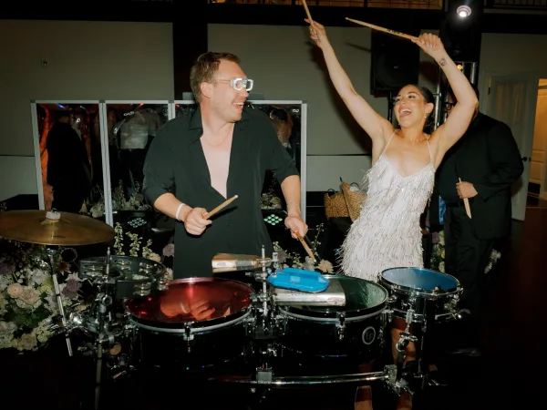 Reception dance moment as bride in a beaded fringe dress holds drumsticks on stage by a drum kit under stage lights at an indoor venue