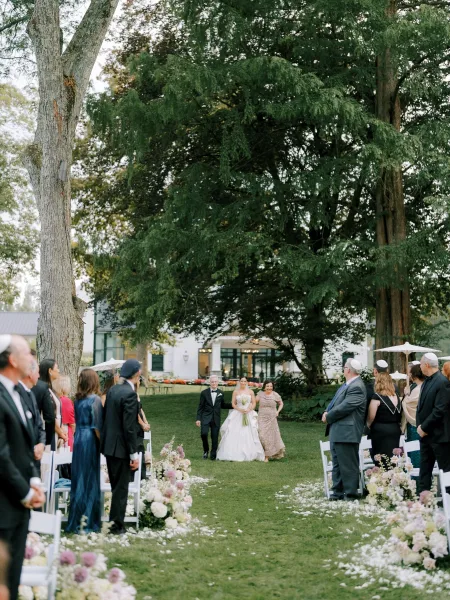 Wedding processional as bride walking down aisle in a strapless ballgown with simple bouquet, escorted past white chairs and petals on a garden lawn