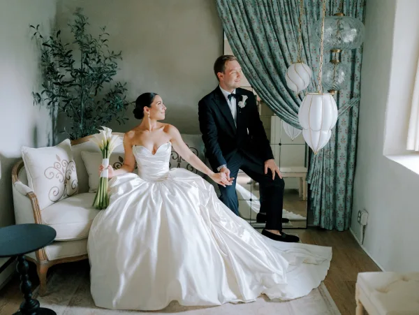 Couple portrait of bride and groom sitting hand in hand on a sofa, her calla lily bouquet and his tuxedo lit by window light
