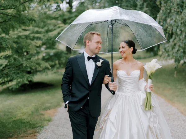 Couple portrait from rain wedding photos, bride and groom walking arm in arm under a clear umbrella on a tree-lined path, bouquet of calla lilies