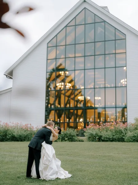 Wedding kiss portrait of bride and groom kissing, bride in strapless satin dress, on lawn outside modern glass barn with chandelier lights at twilight