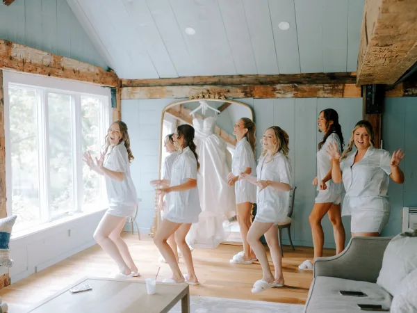 Bridesmaids getting ready in bridal party pajamas, laughing in matching sets as the wedding dress hangs by a bright window in a rustic suite