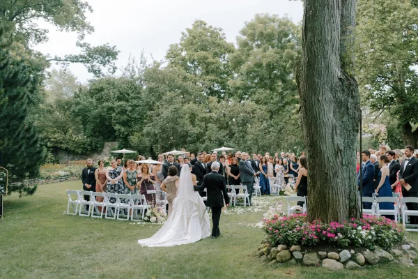 Wedding processional as bride walking down aisle in a strapless ballgown with cathedral veil, guests standing by petals on a garden lawn