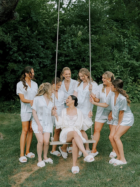 Bridesmaid getting ready in bridal party pajamas, wearing a satin pajama set and slippers beside a rope swing on a green lawn.
