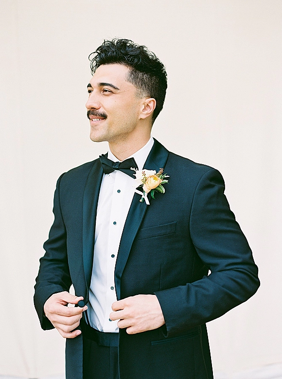 Groom portrait in a black tuxedo and bow tie, wearing a rose boutonniere, posed against a neutral wall in classic black-tie style