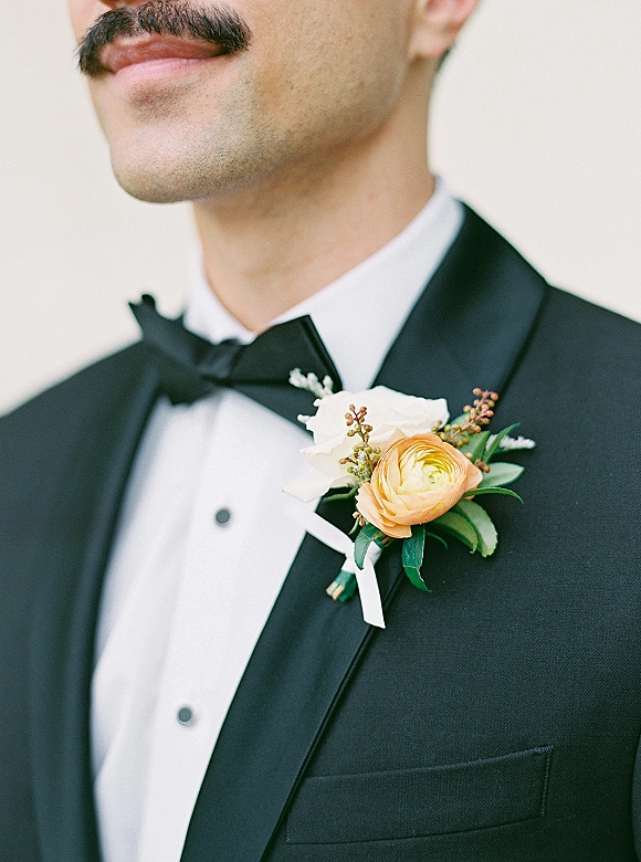 Groom boutonniere with ranunculus boutonniere and white rose pinned to a black tuxedo lapel, ribbon-wrapped with greenery against a neutral wall