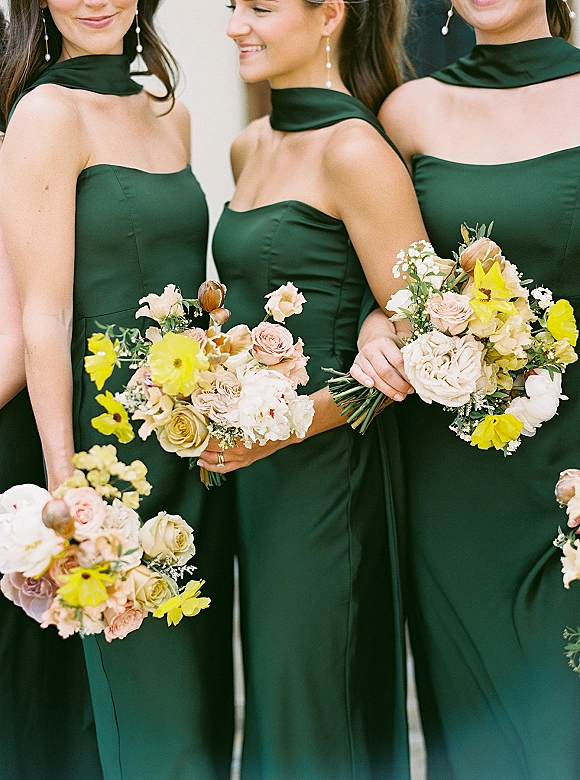 Bridesmaid portrait in dark green bridesmaid dresses with halter scarves, holding rose and yellow bouquets against a light indoor wall