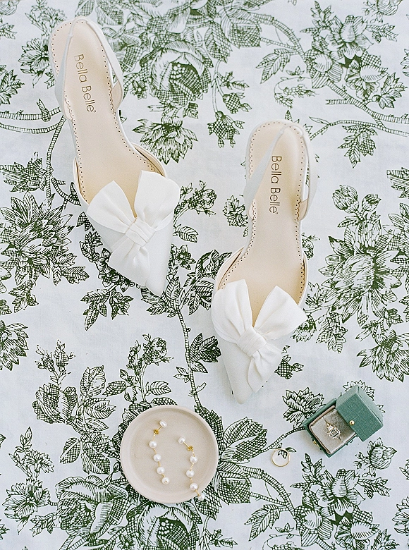 Bridal shoes with white bow heels in a flat lay beside pearl drop earrings and an engagement ring on green floral fabric backdrop