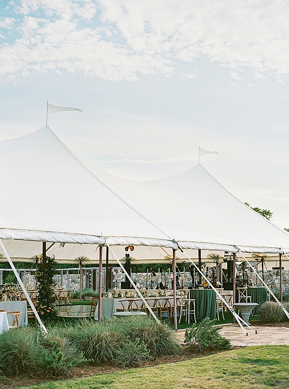 Wedding reception tent with sailcloth canopy, string lights, round tables and cross back chairs on a lawn beside a stone wall