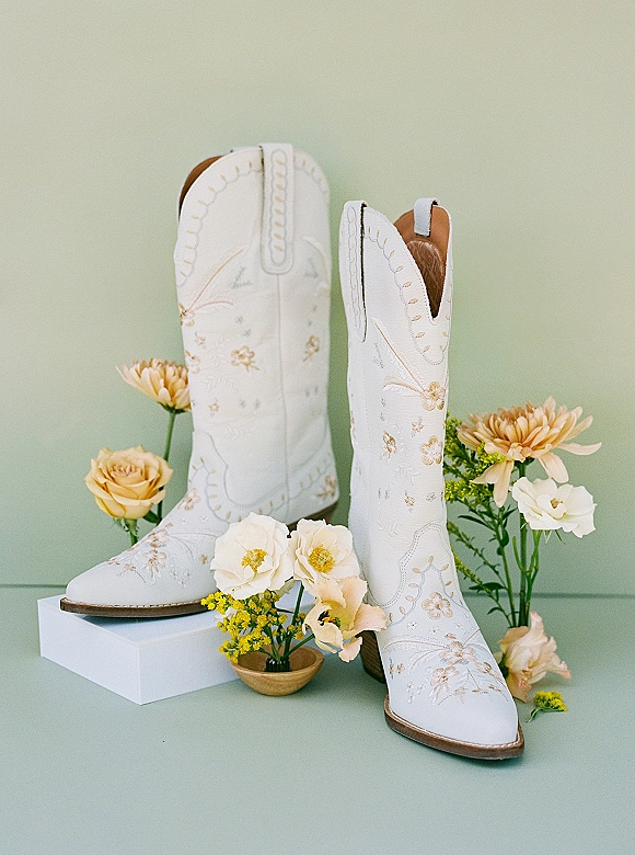 Bridal cowboy boots in white leather with floral embroidery, styled beside wedding flowers on a tabletop against a sage green backdrop