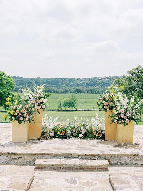 Ceremony altar decor with rose and greenery floral arrangements on wooden pedestal stands and ground florals on a stone patio with rolling hills beyond