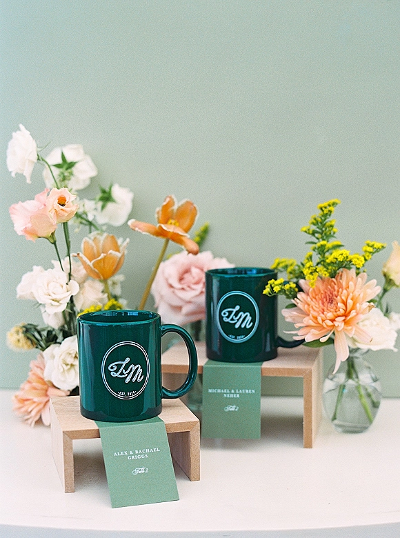 Wedding favors displayed as custom wedding mugs with place cards and bud vases on wooden risers against a pastel green backdrop